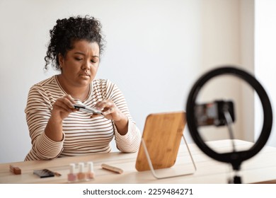 Unhappy young hispanic woman plus size beauty blogger recording video for followers, sitting at desk full of cosmetics, holding eyeshadow palette, streaming from home, using blogger set, copy space - Powered by Shutterstock