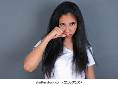 Unhappy Young hispanic  woman wearing white T-shirt over grey background crying while posing at camera whipping tears with hand.
