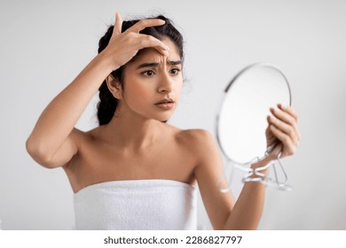 Unhappy Young Indian Woman Looking In Mirror And Checking Wrinkles On Her Face, Attractive Estern Female Standing Examining Skin Fine Lines While Standing Wrapped In Towel After Bath, Closeup - Shutterstock ID 2286827797