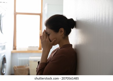 Unhappy young Indian woman standing in domestic laundry room and cover face with palms crying feels miserable. Family problems, break up, marriage split, resentment against her husband, abuse concept