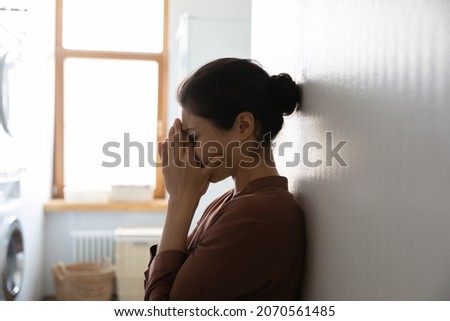 Unhappy young Indian woman standing in domestic laundry room and cover face with palms crying feels miserable. Family problems, break up, marriage split, resentment against her husband, abuse concept