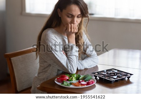 Unhappy young Latin woman look at chocolates and vegetables face temptation suffer from eating disorder. Millennial female think of healthy food choice, diet. Wellbeing, weight loss concept.