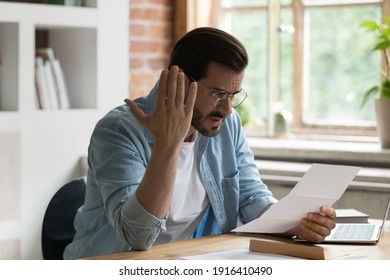 Unhappy Young Male Employee Feel Stressed Read Bad News In Postal Paper Letter At Workplace. Upset Angry Caucasian Man Worker Distressed With Negative Message In Post Paperwork Or Document.