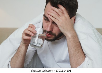 Unhappy Young Man With Covid19 Or Flu Touching His Forehead And Drinking Water From Glass While Sitting On Bed During Self Isolation At Home