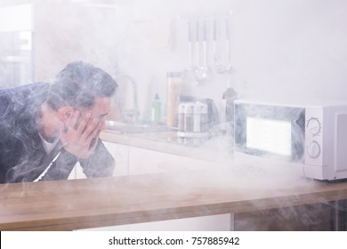 Unhappy Young Man Looking At Fire Coming From Microwave Oven In Kitchen