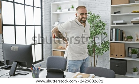 Unhappy young man worker suffering from backache while standing indoors at the office, serious business professional touching injured spinal column, worried about painful back injury at workplace