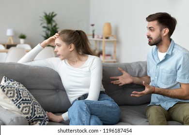 Unhappy Young Millennial Married Couple Sitting On Couch In Living Room At Home And Quarrelling. Husband Arguing Emotionally Talking Frustrated Wife Looking Away. Break Up And Misunderstanding Concept