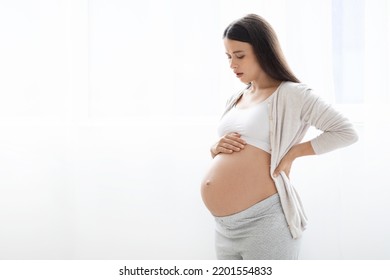 Unhappy Young Pregnant Woman In Pajamas Having Back Pain, Standing Next To Window At Home, Touching Ache Zone And Big Tummy, Suffering From Backache, Panorama With Copy Space