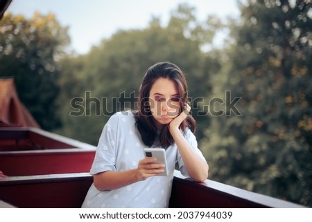 Unhappy Young Woman Checking her Smartphone in the Morning. Sad bored adult person sitting in the balcony using her mobile phone as distraction

