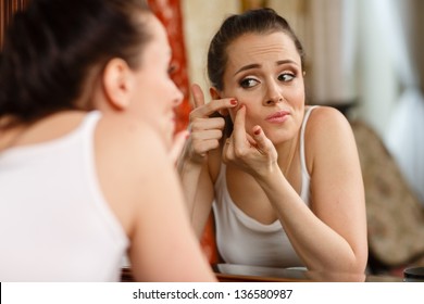 Unhappy young woman finding an acne on a cheek, sits before a mirror.
