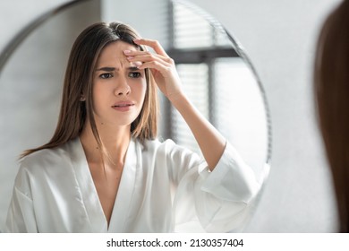 Unhappy Young Woman Looking In Mirror And Touching Wrinkles On Her Face, Attractive Millennial Female Standing In Bathroom And Examining Skin Fine Lines, Selective Focus On Reflection, Closeup