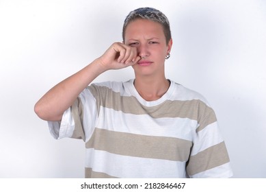 Unhappy young woman with short hair wearing striped t-shirt over white background crying while posing at camera whipping tears with hand.