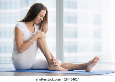 Unhappy young woman sitting on the mat, grabbing an ankle, unable to start yoga work out because of sport injury, feeling pain. Beginner doing wrong exercise without coacher