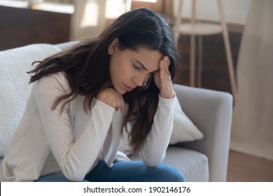 Unhappy young woman touching forehead, yearning, thinking about relationship problems, feeling depressed and lonely, sitting on couch alone, frustrated female suffering from headache or migraine