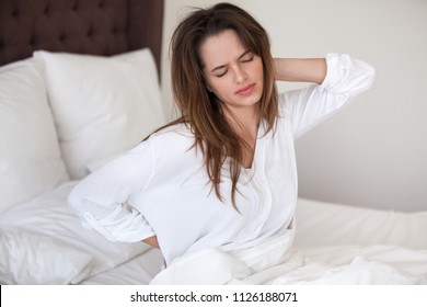 Unhappy young woman waking up in uncomfortable bed feeling ache in back pain massaging tensed muscles of stiff neck after sleep on bad mattress in incorrect posture, fibromyalgia and backache concept