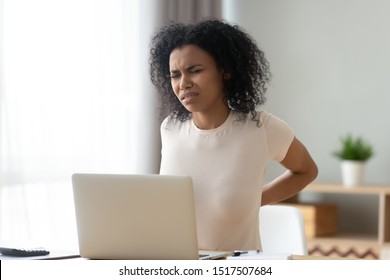 Unhealthy african woman need treatment for lower back pain caused by computer long usage overuse, frown female sitting at desk touch massaging dorsum to reducing ache unpleasant discomfort sensations