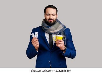 Unhealthy bearded man standing and holding yellow cup with white tissue, drink hot tea, has flu or influenza, wearing official style suit. Indoor studio shot isolated on gray background.