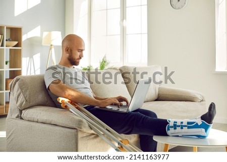 Unhealthy businessman with injured leg in bandage sit on sofa at home work online on computer. Unwell male employee with foot trauma wearing special splint use laptop. Injury and rehabilitation.