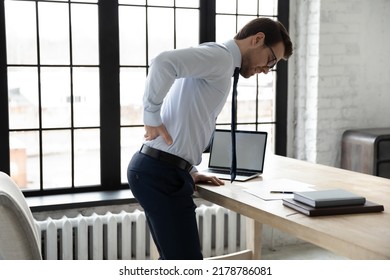 Unhealthy Businessman Wearing Glasses Touching Lower Back, Suffering From Backache, Standing Near Desk In Office, Stressed Employee Feeling Unhealthy And Unwell After Long Hours Sedentary Work