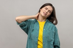 Unhealthy Dark Haired Woman Touching Neck Feeling Pain And Numbness, Worried About Muscle Tension, Osteochondrosis, Wearing Casual Style Jacket. Indoor Studio Shot Isolated On Gray Background.