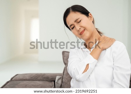 Unhealthy dark haired woman touching neck feeling pain and numbness, worried about muscle tension, osteochondrosis, wearing casual style shirt. Indoor living room isolated on white wall background.