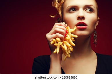 Unhealthy eating. Junk food concept. Portrait of fashionable young woman holding (eating) fried potato (fries, chips) in her hand and posing over red background. Close up. Copy-space. Studio shot