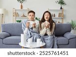 Unhealthy family couple sitting on the sofa, wrapped in blankets, feeling flu or cold symptoms during winter season. Young man and woman in warm clothes blowing noses and sneezing in paper towels.