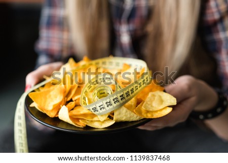 unhealthy fast food snacks. weight gain due to bad nutrition habits. woman hands holding chips with a measure tape on a plate