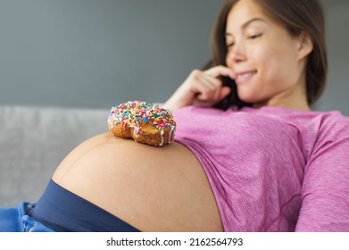 Unhealthy Food During Pregnancy. Sugar Temptation Craving Asian Pregnant Woman Trying To Resist Eating A Sweet Donut On Her Baby Belly. Funny Eating Cravings Concept