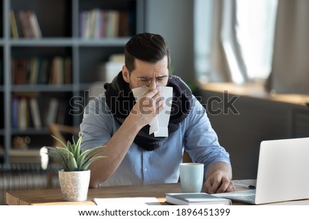 Unhealthy ill stressed young 30s businessman sitting at table covered in warm scarf, using paper tissue suffering from runny nose, having first flu grippe symptoms, seasonal disease concept.