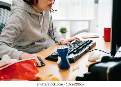 Unhealthy Lifestyle. Close Up Of Young Funny College Student In Summer Break Wearing Hoodie Hands Playing Online Games With Mouse And Keyboard Talking With Friends In Headphones Eating Junk Food.