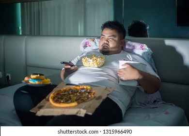 Unhealthy lifestyle concept: Asian obese man eating junk foods while watching TV in bed before sleep
