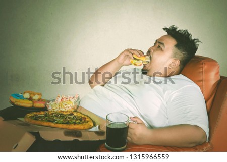 Unhealthy lifestyle concept: Young fat man eating burger and junk foods with gluttony expression while watching TV on the sofa
