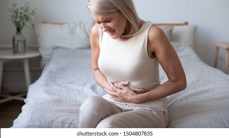 Unhealthy mature woman holding belly, feeling discomfort, health problem concept, unhappy older female sitting on bed, suffering from stomachache, food poisoning, gastritis, abdominal pain, climax