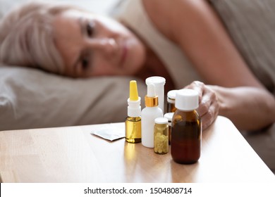 Unhealthy Mature Woman Lying In Bed, Taking Medicine From Cold Or Insomnia, Unhappy Older Female Suffering From Flu Or Seasonal Fever, Treatment Concept, Pills And Meds On Bedside Table Close Up