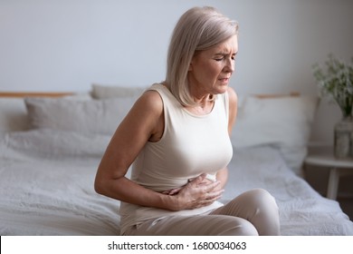 Unhealthy Middle Aged Frowning Woman Sitting On Bed, Putting Hands On Belly, Suffering From Strong Stomach Ache. Unhappy Older Lady Having Painful Feeling In Abdomen, Pancreatitis Gastritis Symptom.