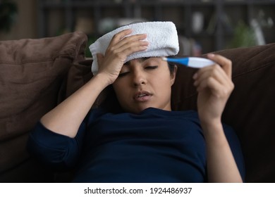 Unhealthy millennial Indian woman struggle with high temperature measure with thermometer. Sick ill young ethnic female suffer from flu or cold, have covid-19 lying at home. Corona virus concept.