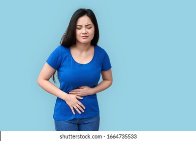 Unhealthy And Sad Young Adult Asian Girl Standing Isolated On Grey Background With Spasm In Stomach, Holding Hands On Belly