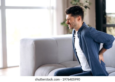 Unhealthy Sad Young Adult Man Stand Up From Couch In Living Room Touches Back, Suffer From Back Pain. Caucasian Guy Suffers From Arthritis, Radiculitis, Backache Pain