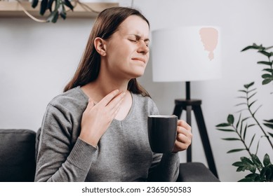 Unhealthy sad young girl touch neck hard to swallow having discomfort or painful feeling, unwell caucasian woman holding cup suffering from angina or tonsillitis, sore throat, experience loss of voice