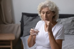 Unhealthy Senior Woman Wake Up Sits On Bed In Bedroom Holding Glass Of Water Take Pill Suffer From Migraine Relieve Pain, Treatment Prevent Insomnia Sleep Disorder, Senile Diseases Prevention Concept