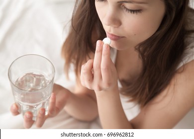Unhealthy sick woman suffers from insomnia or headache, takes sleeping pill while sitting in bed with glass of water, depressed girl holds antidepressant meds, painkiller for menstrual pain, close up