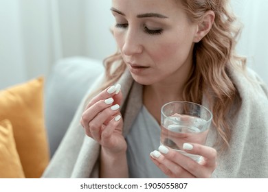 Unhealthy Sick Young Woman Covered In A Plaid, Takes Sleeping Pill From Insomnia Or Headache. Female With Glass Of Water, Holds Antidepressant Meds Or Painkiller For Menstrual Pain