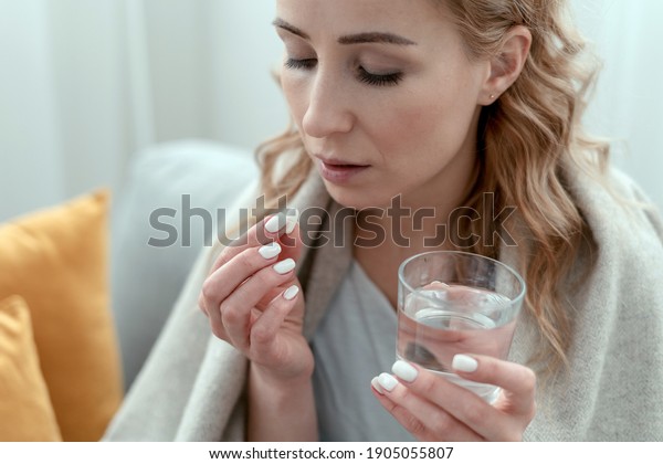 Unhealthy sick young woman covered in a plaid, takes\
sleeping pill from insomnia or headache. Female with glass of\
water, holds antidepressant meds or painkiller for menstrual\
pain