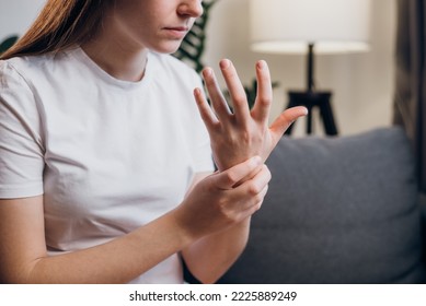 Unhealthy Upset Young Caucasian Woman With Wrist Pain Tries To Relieve Suffering Sitting On Sofa In Living Room At Home. Close-up Of Wrist Female Hand With Arthritic Joints. Wrist Pain Concept.