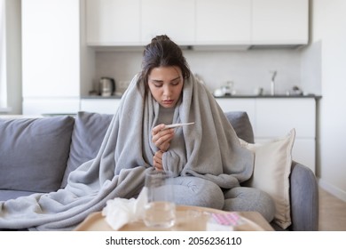 Unhealthy upset young female with fever looking at thermometer with high degree temperature. Sick ill girl suffer from flu or cold, have covid-19 symptoms sitting covered blanket on couch at home. - Shutterstock ID 2056236110