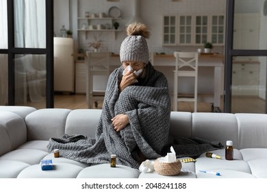 Unhealthy woman sit in blanket hat feel sick suffer from flu or covid-19 corona virus. Unwell young Caucasian female sneeze sniffle have running nose, struggle with influenza or cold at home. - Shutterstock ID 2048241188