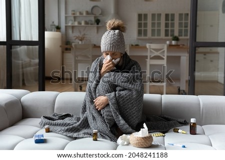 Unhealthy woman sit in blanket hat feel sick suffer from flu or covid-19 corona virus. Unwell young Caucasian female sneeze sniffle have running nose, struggle with influenza or cold at home.