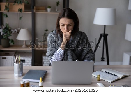 Unhealthy young Caucasian woman sit at desk at home office work online on computer suffer from covid-19 virus. Sick female busy at laptop cough struggle with coronavirus or flu at workplace.