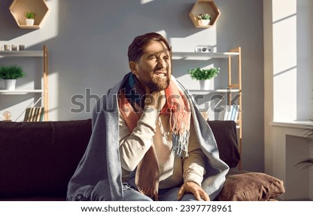 Unhealthy young man feels severe pain and sore throat, malaise and weakness. Bearded man is sitting at home in living room, wrapped in warm blanket, dressed in sweater and jeans. Healthcare concept.